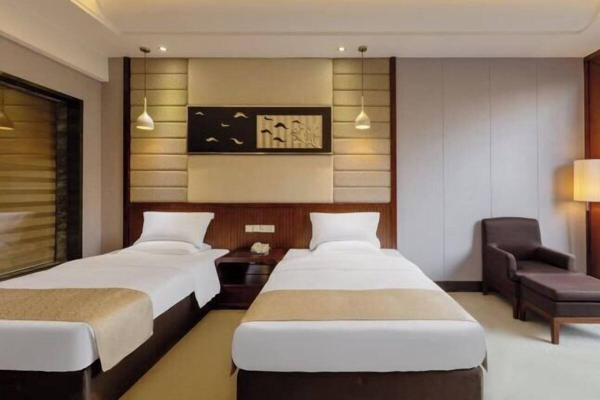 Habitación de hotel con dos camas, cabecero de madera, una obra de arte colgada en la pared y un rincón de descanso con una silla y una mesa.