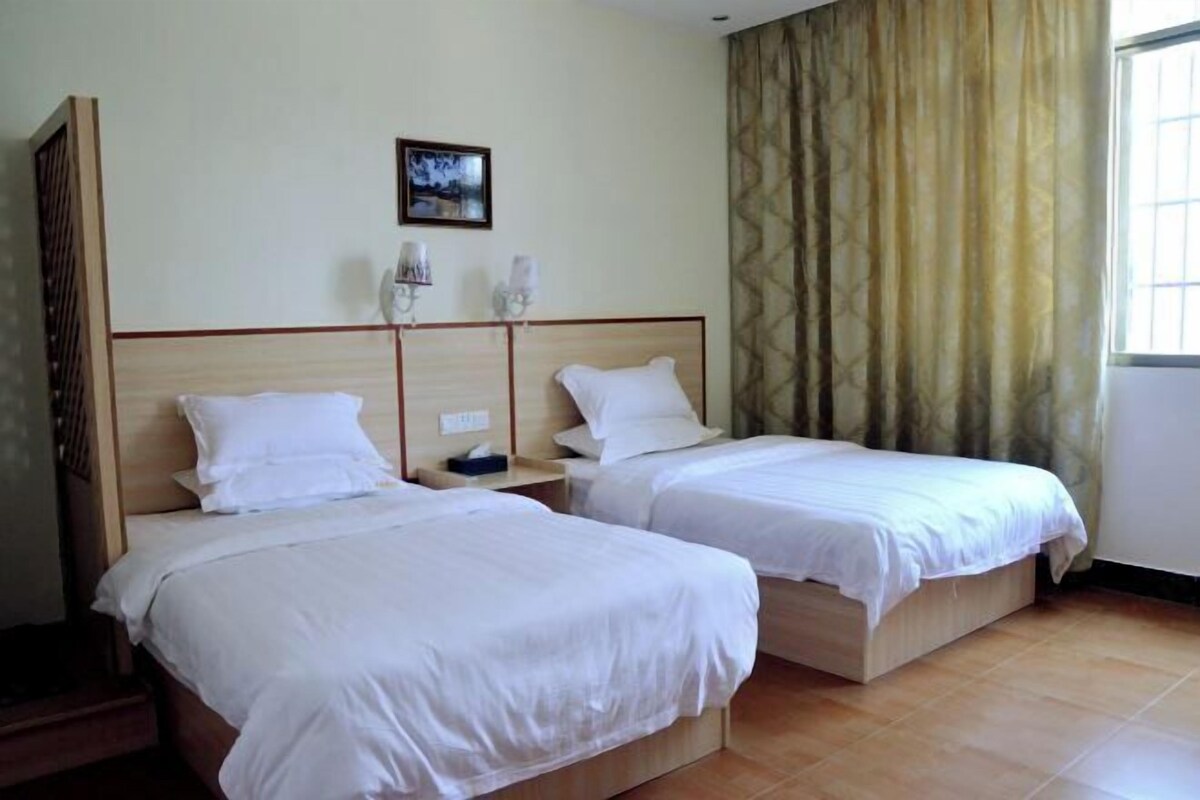 Une chambre d’hôtel avec deux lits simples, une tête de lit en bois, une table de chevet, une lampe fixée au mur et une fenêtre avec des rideaux.