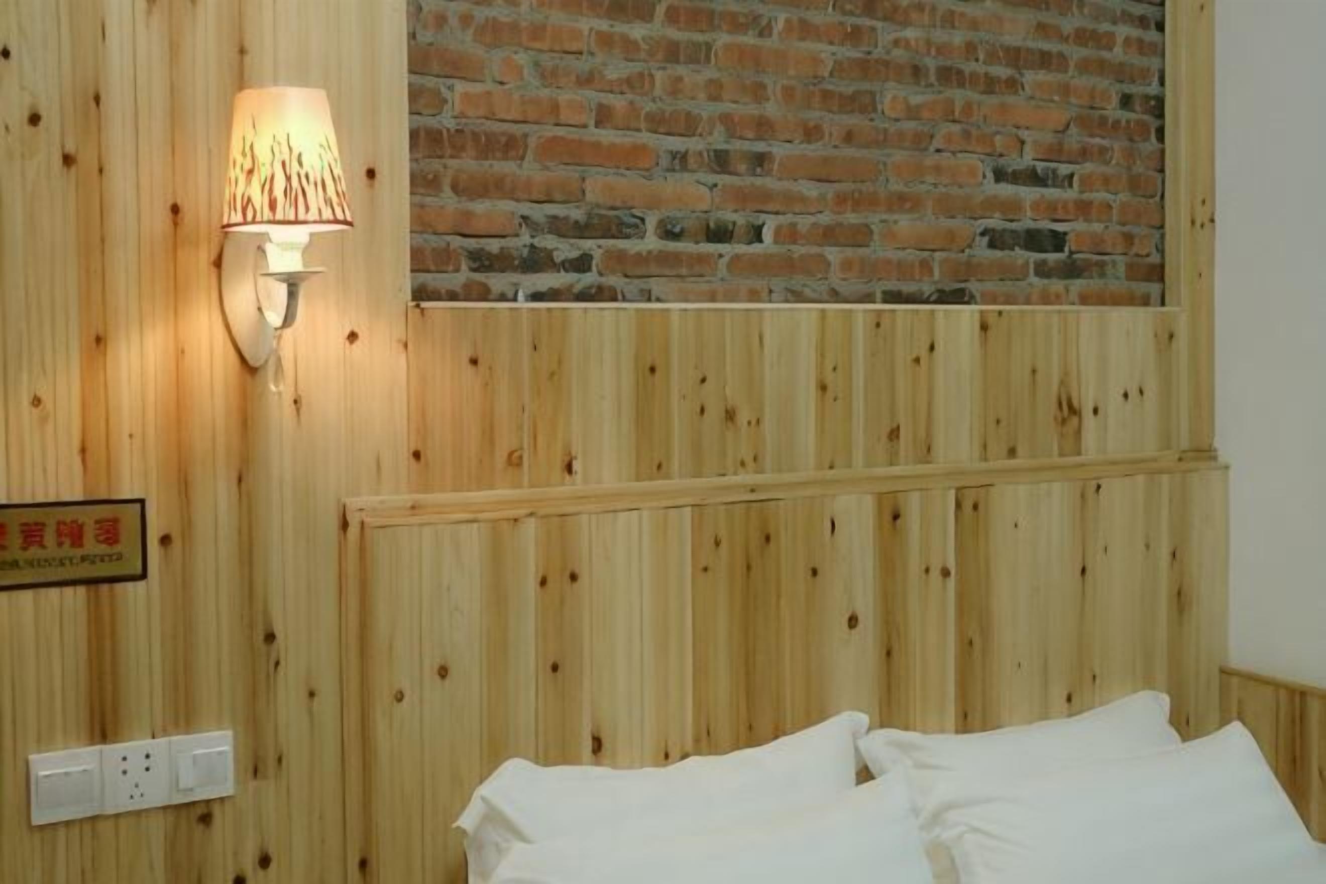 Eine Wand mit Holzverkleidung und einem Ziegelbereich, ein Bett mit weißer Bettwäsche, eine Wandlampe sowie ein Schild mit chinesischen Schriftzeichen.