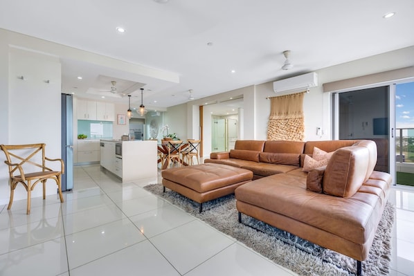 A modern living room with a large brown leather sofa, a dining area with a table and chairs, and a kitchen in the background. - Skytower Wonder Oceanview Balcony - Pool - Office (Darwin)