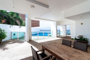 A spacious outdoor area with a wooden table and chairs, a large potted plant, and a view of a swimming pool and ocean. - Skytower Wonder Oceanview Balcony - Pool - Office (Darwin)