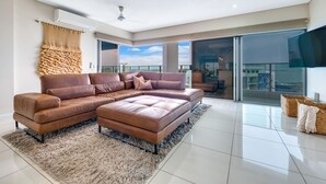 A spacious living room with a large sectional sofa, a fluffy rug, and a large window offering a view of the city. - Skytower Wonder Oceanview Balcony - Pool - Office (Darwin)