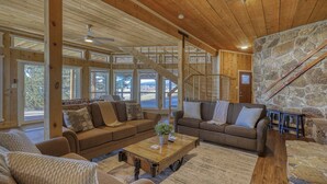 A spacious living room with wooden beams, large windows, a stone wall, and a staircase.
