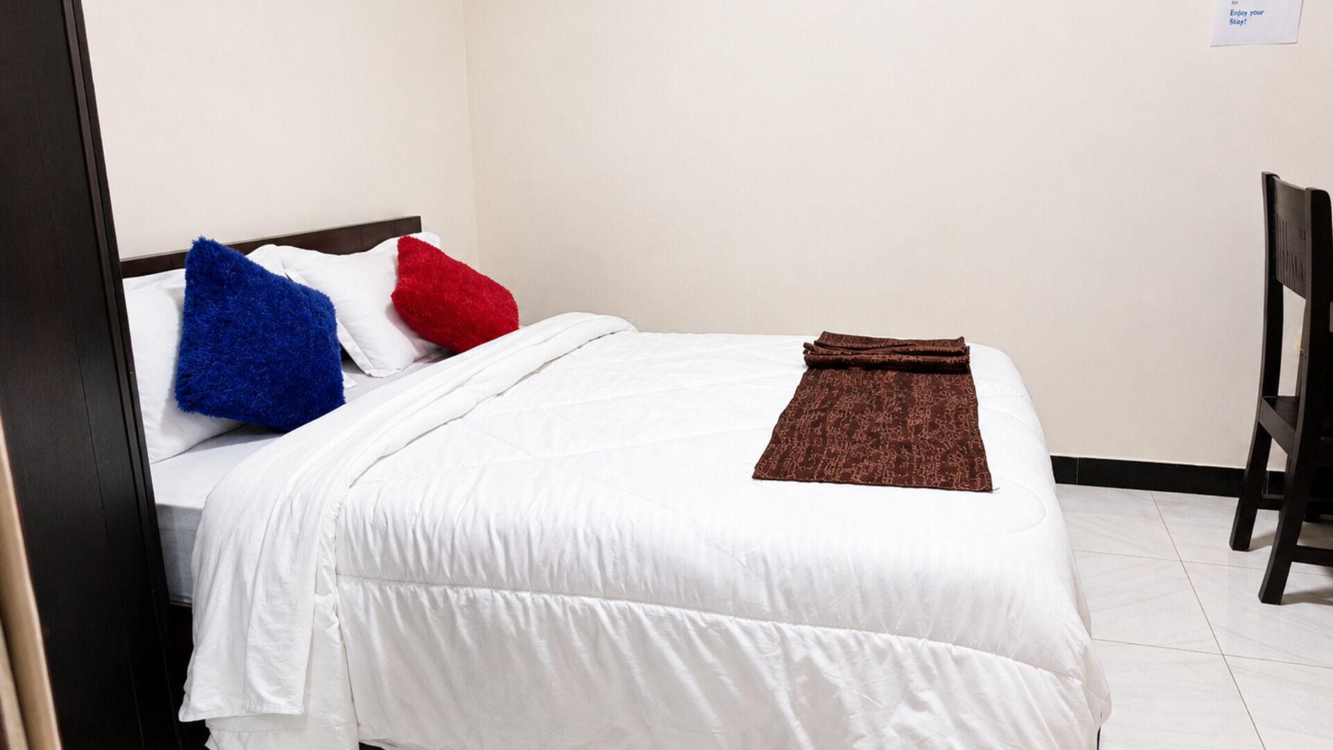 Una cama bien hecha con ropa blanca, una almohada azul y roja, y un plaid marrón.