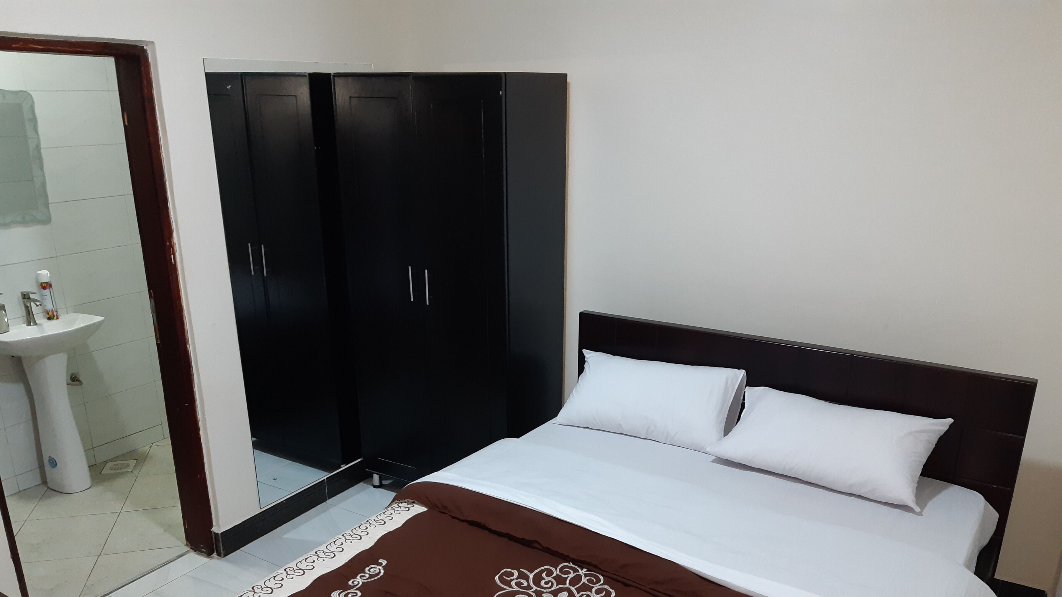 Quarto com cama, travesseiros brancos, colcha marrom, guarda-roupa preto e pia branca.