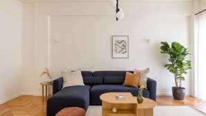 A modern living room with a dark blue sofa, a wooden coffee table, a small side table, a large potted plant, and a framed abstract artwork on the wall. - Green Oasis 2BR Apartment in Pangrati (Athina)