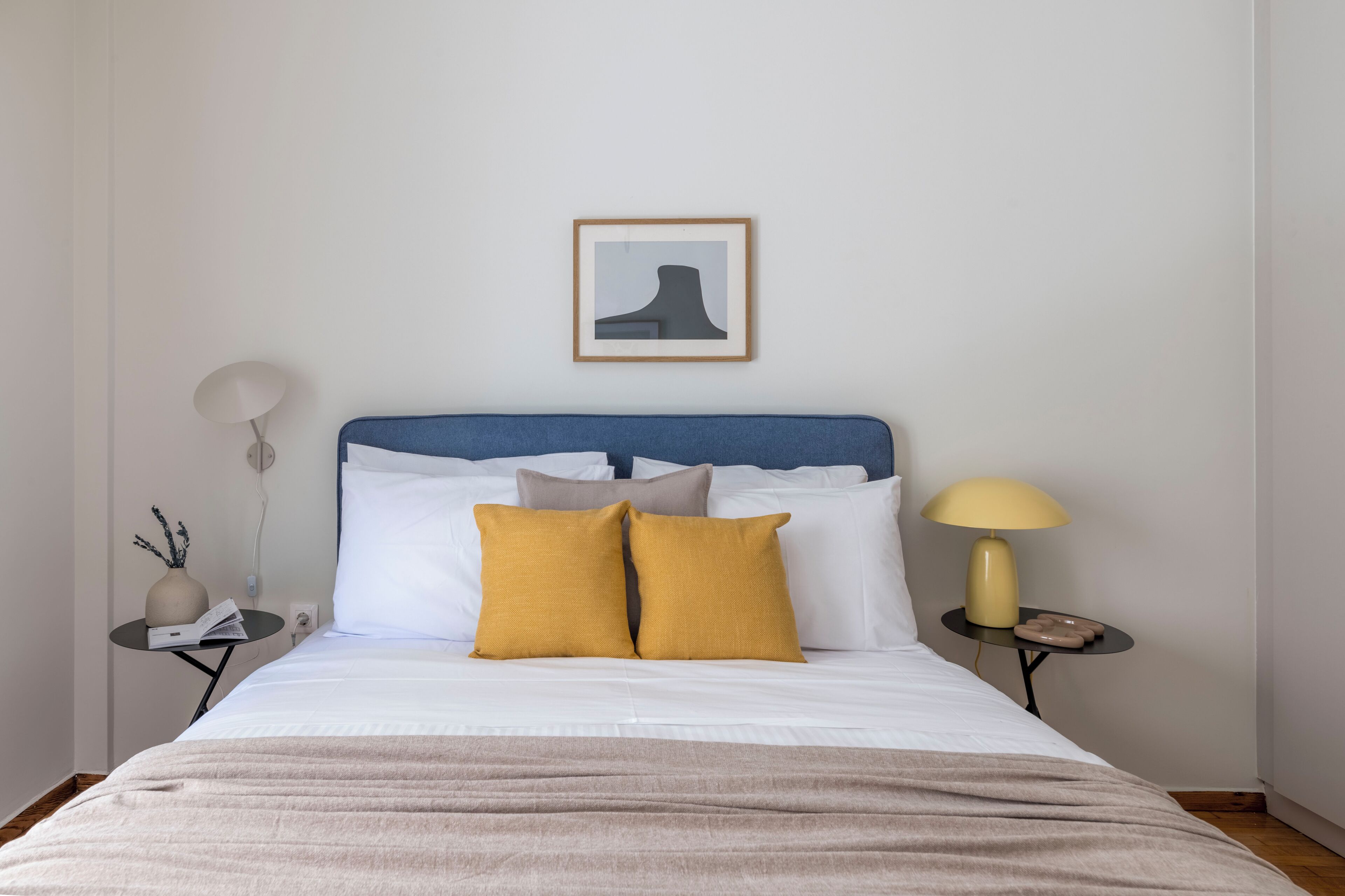 A neatly made bed with a blue headboard, white bedding, and two mustard pillows. There is a framed picture on the wall above the bed, and two bedside tables with lamps and decorative items.