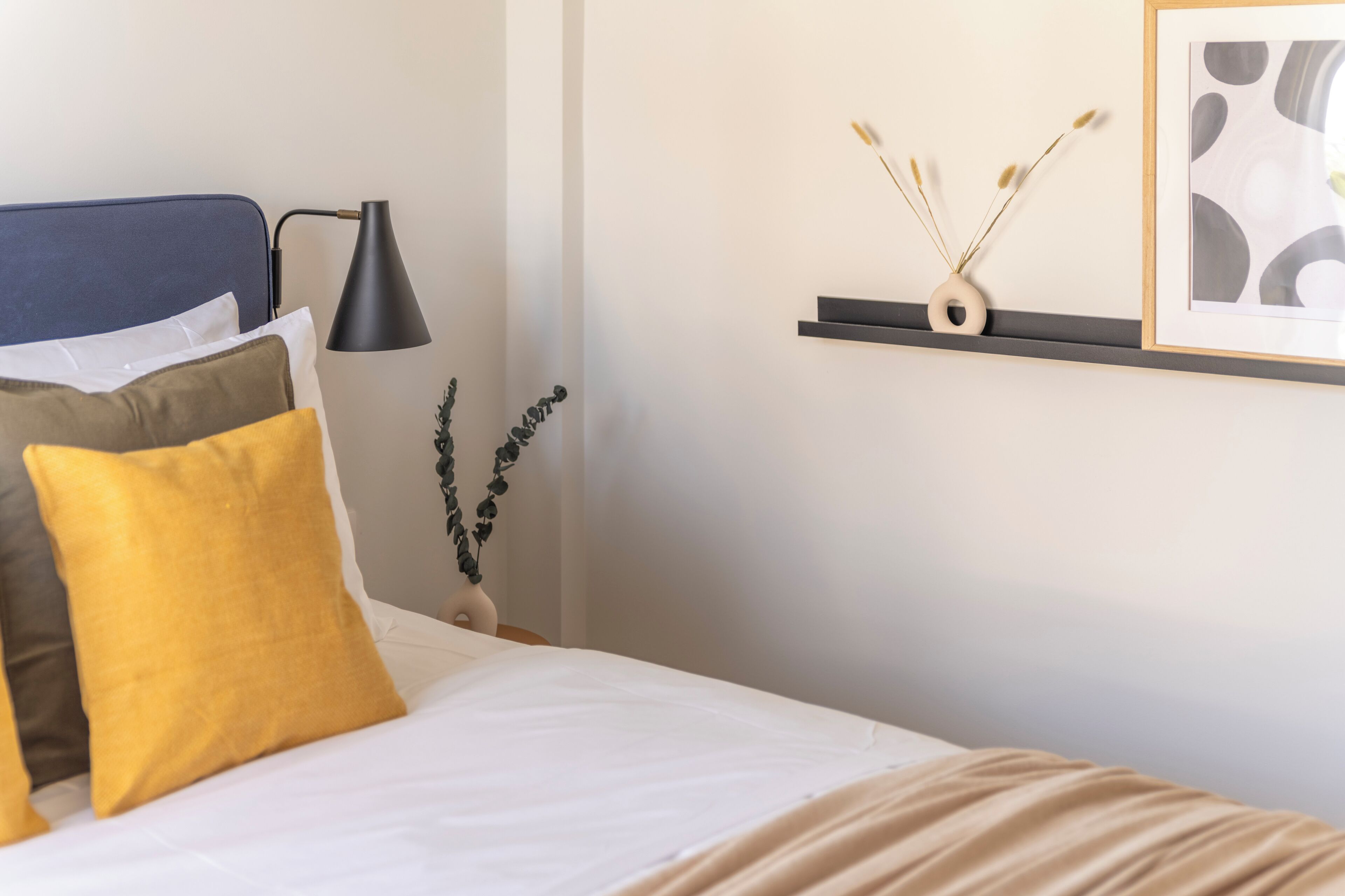 A neatly made bed with white and yellow pillows, a black lamp, and a wall-mounted shelf with decorative items.