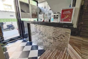 Uma área de recepção com piso xadrez, parede de tijolos e um balcão com diversos avisos e um cartaz.