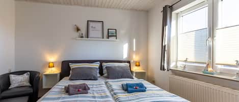 A bedroom with two beds, bedside lamps, a nightstand, a chair, and a window with blinds.