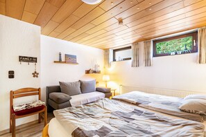 Une pièce avec un plafond en bois, un lit avec une literie à motifs, un canapé, une chaise, une petite table, une lampe, une fenêtre avec des rideaux et une étagère fixée au mur, sur laquelle se trouvent une petite statue et un tableau encadré.