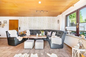 Un salon moderne avec un canapé d’angle, un plafond en bois, une table à manger et une fenêtre avec des stores.