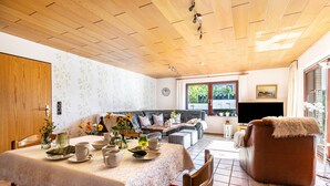 A dining area with a table set for four, a sofa, and a television in a room with wooden ceiling and large windows.