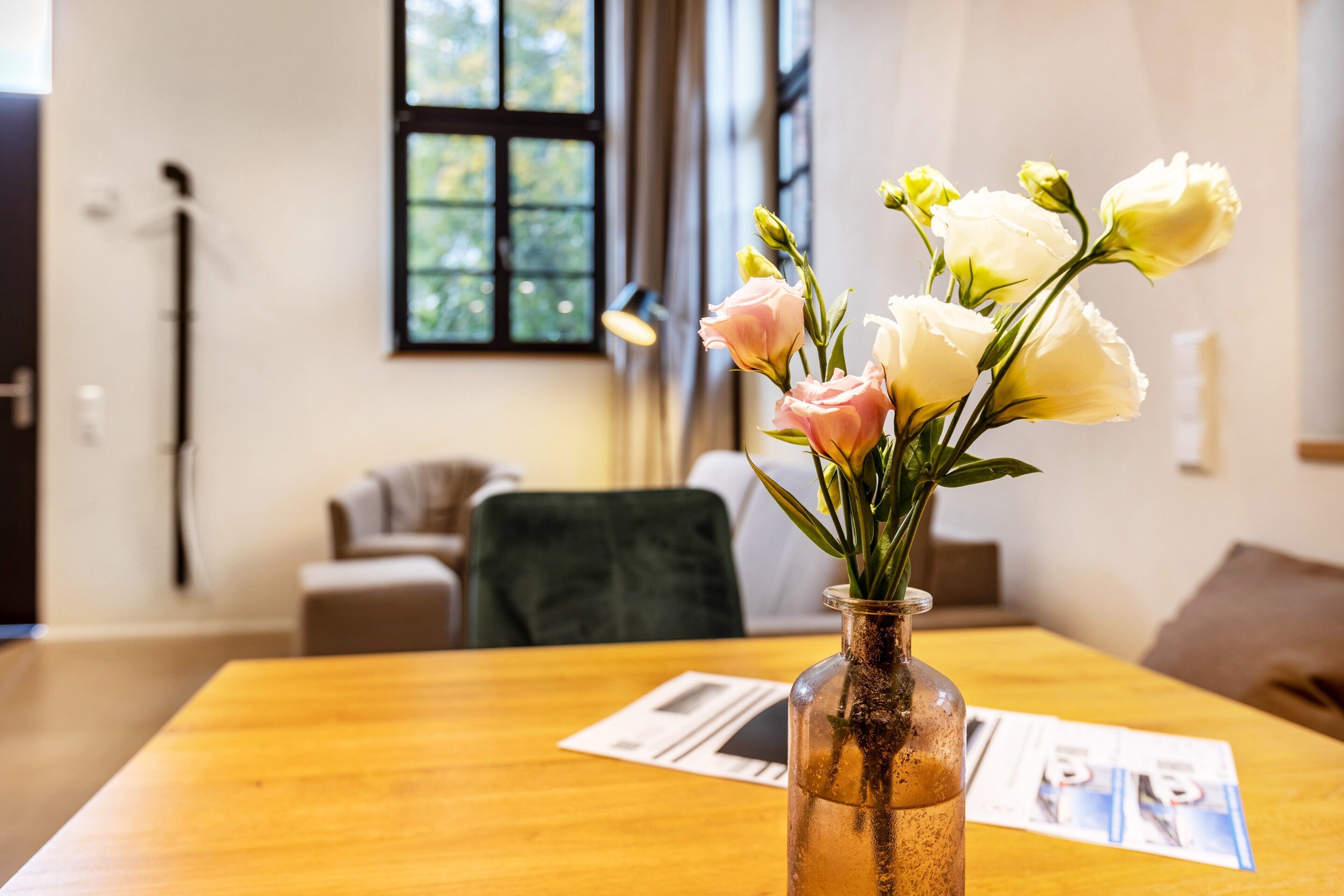 Ein Holztisch mit Blumen in einer Vase, einer Lampe und einem Fenster im Hintergrund.