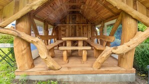 Een houten constructie met een centrale tafel en banken, omringd door grote boomstammen.