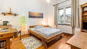 Een houten bed met hoofdeinde, een nachtkastje, een lamp, een raam met gordijnen, een plank met decoratieve objecten en een schilderij aan de muur.