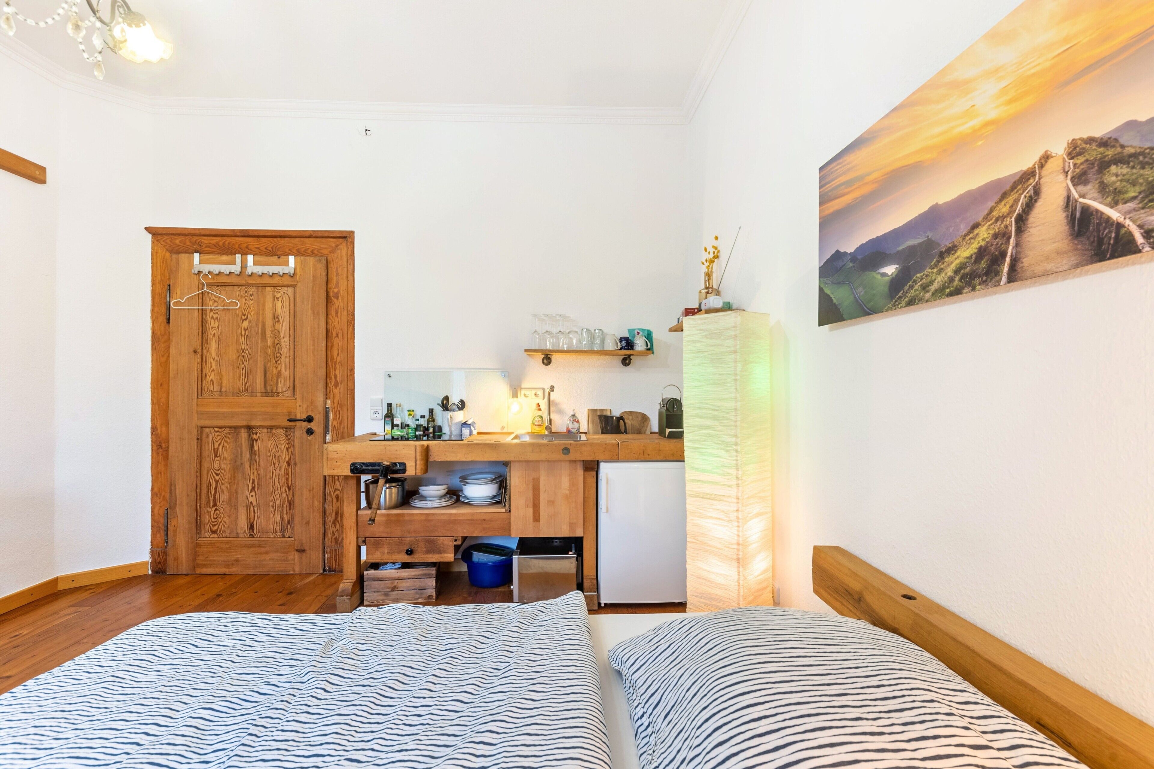 Ein kleines Zimmer mit Holztür, einer Küchenzeile, einem Bett mit gestreifter Bettwäsche und einem Landschaftsbild.
