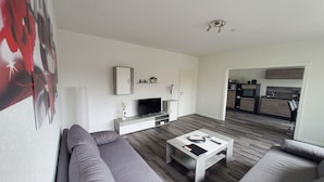 Una sala de estar moderna con un sofá gris, una mesa de centro, un televisor y una cocina al fondo.