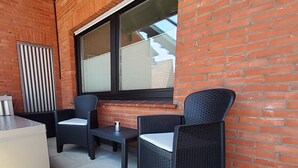 Un patio con dos sillones de mimbre, una mesa pequeña, un radiador de metal y una ventana con persianas.