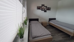 Una habitación pequeña y limpia con dos camas individuales, una mesita de noche, una planta en maceta y una obra de arte colgada en la pared.