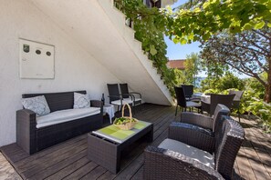 Un patio exterior con muebles de mimbre, una mesita con una cesta y una pared blanca con un ventanal.