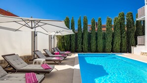 A poolside area with lounge chairs and umbrellas, surrounded by tall trees and a clear blue sky. - House Rose With Private Pool And Garden (Krk)