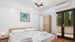 A modern bedroom with a large bed, wooden headboard, a bedside table with a lamp, a painting on the wall, and a balcony with a view of trees. - House Rose With Private Pool And Garden (Krk)