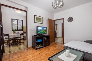 A hotel room with a wooden floor, a bed, a TV, a dining area with a table and chairs, and a door leading to another room. - House Rose With Private Pool And Garden (Krk)