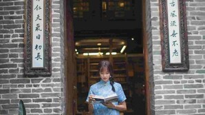 Une personne vêtue d’une robe bleue, debout dans un doorway, avec un mur de briques et une enseigne portant des caractères chinois.
