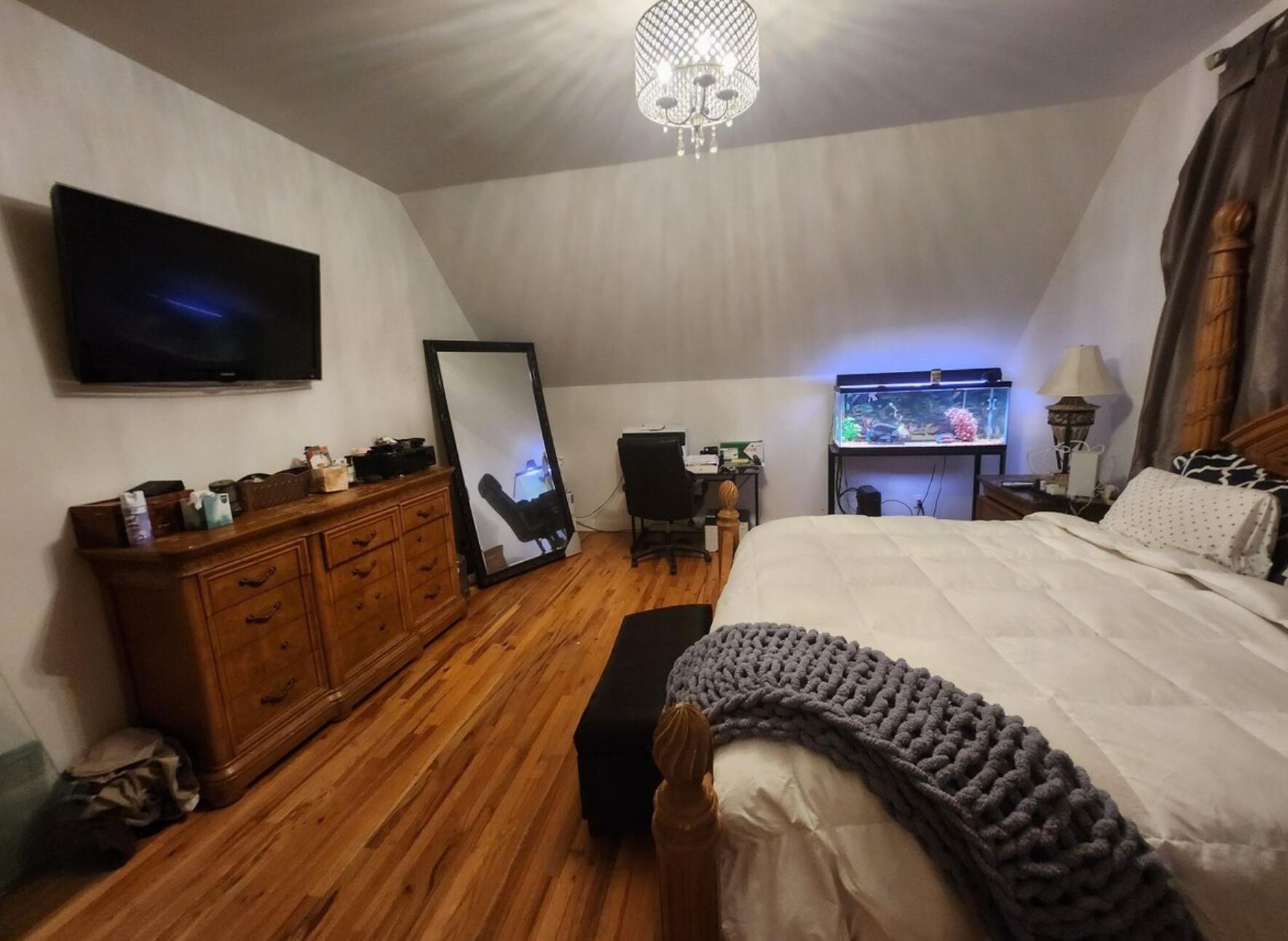Un dormitorio con una cama grande, cómoda de madera, televisor, espejo, escritorio con acuario y una araña.
