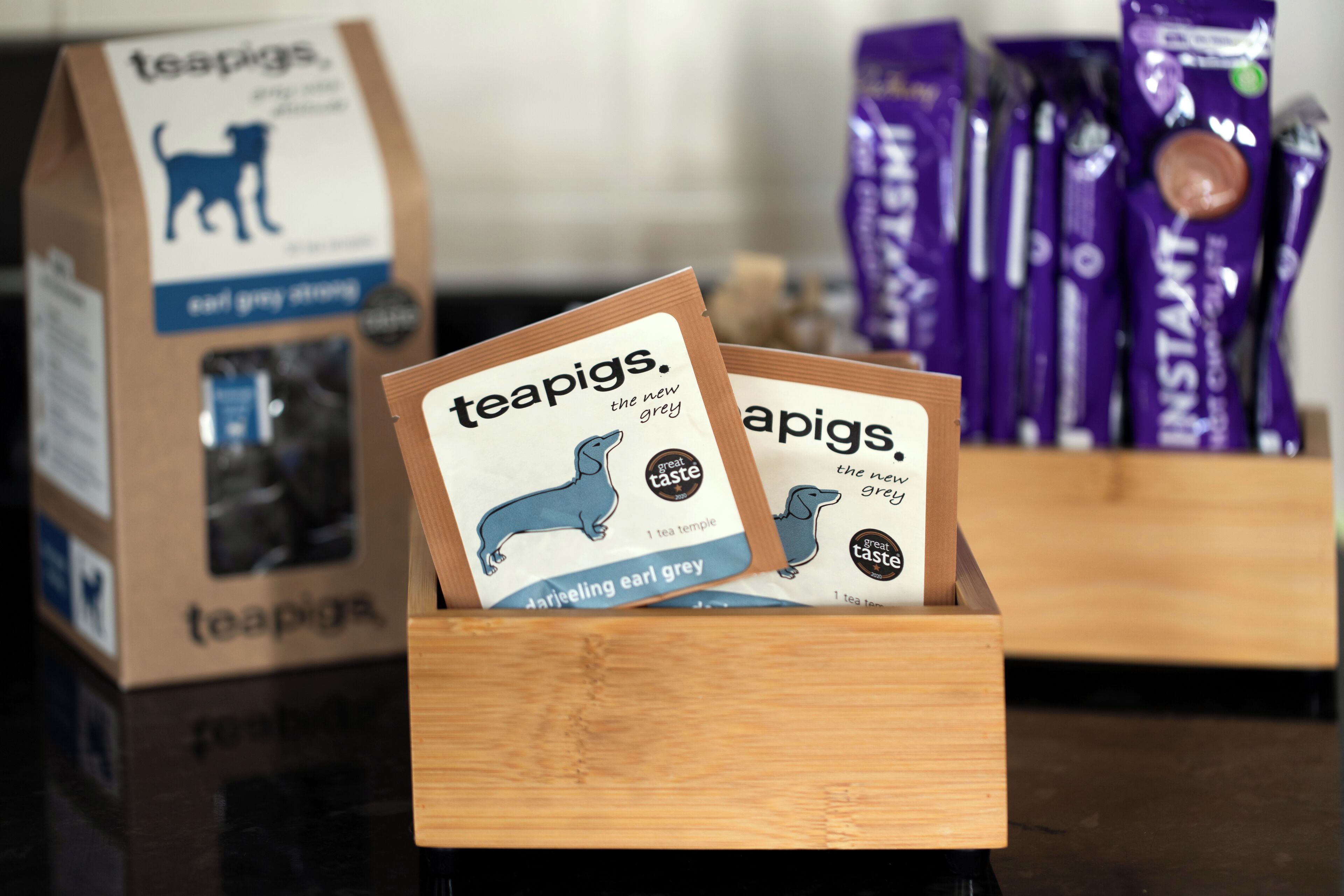 Une boîte en bois contenant des sachets de thé étiquetés « teapigs. the new grey ». En arrière-plan, on aperçoit d’autres sachets de thé et une boîte en carton ornée d’une illustration de chien.