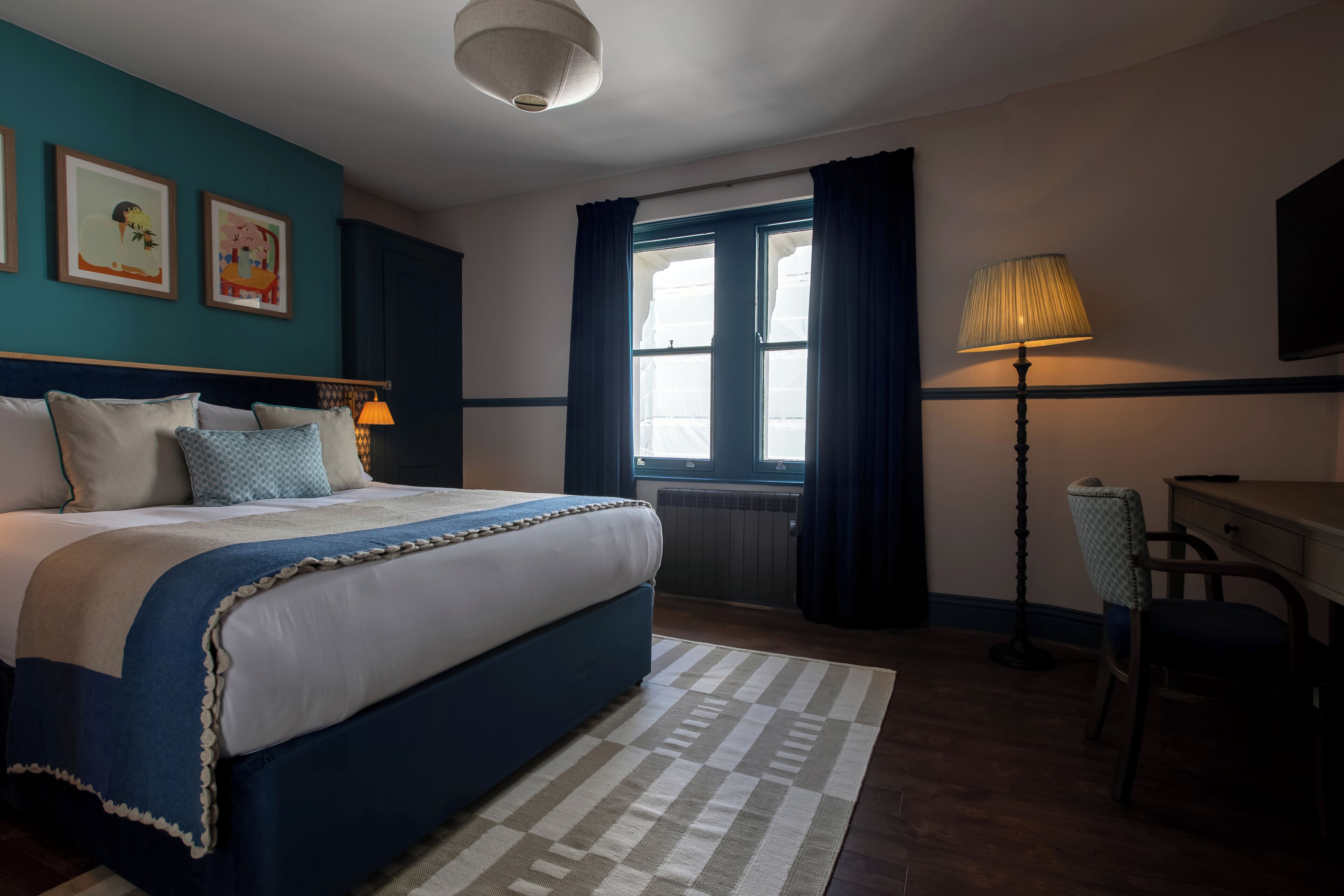 A neatly arranged bedroom with a large bed, a desk, and a chair. There is a window with curtains, a lamp, and framed pictures on the wall.