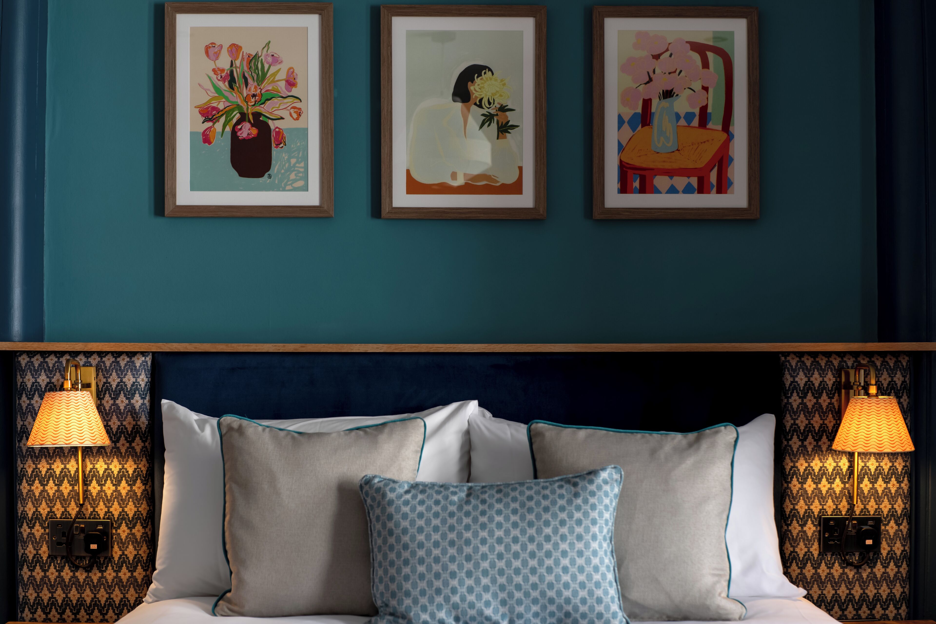 A hotel room with a teal wall, a bed with white and blue patterned pillows, and three framed artworks above the bed.