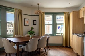 Un comedor con una mesa de madera y seis sillas, una lámpara colgante y vistas a edificios a través de ventanas amplias con cortinas.