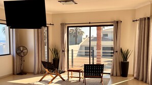 Une pièce spacieuse dotée d’une porte-fenêtre coulissante donnant sur un balcon, de deux chaises, d’une petite table, d’un ventilateur sur pied et de deux plantes en pot.