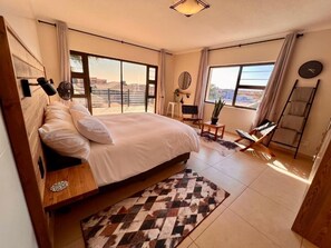 A bedroom with a large bed, a wooden headboard, a small table, a chair, a standing fan, a ladder, and a view of the city through large windows. - Spacious 6-bedroom house, full equipped, in wonderful Swakopmund with WiFi (Swakopmund)