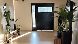 Un hall d’entrée spacieux et lumineux, avec un sol carrelé, une porte noire et plusieurs plantes en pot.