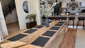 Une longue table de salle à manger en bois, surmontée de sets de table noirs, un vase avec des fleurs blanches et, au fond, une cuisine.