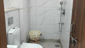 A small, compact bathroom with white marble tiles, a toilet, a shower, and a waste bin. - The Park Ambanc-New Delhi (New Delhi)
