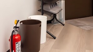 Un coin de pièce avec un mur blanc, une porte marron, un extincteur, une poubelle blanche et une étagère métallique contenant des assiettes.