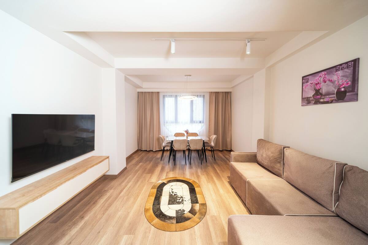Ein modernes Wohnzimmer mit Flachbildfernseher, einer Couch, einem Esstisch mit Stühlen und einem großen Fenster mit Vorhängen.