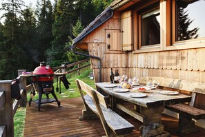 Una terraza de madera con una mesa de picnic preparada para comer, una parrilla y una cabaña con ventanas amplias.