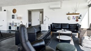 Ein modernes Wohnzimmer mit einem schwarzen Ledersofa, einem runden Couchtisch und einem Korbstuhl. Es gibt eine weiße Kommode und ein kleines Regal mit Dekoration.
