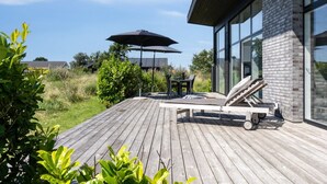 Eine Holzterrasse mit Liegestuhl und Sonnenschirm, ein Backsteinhaus mit großen Fenstern und eine Rasenfläche mit Bäumen.