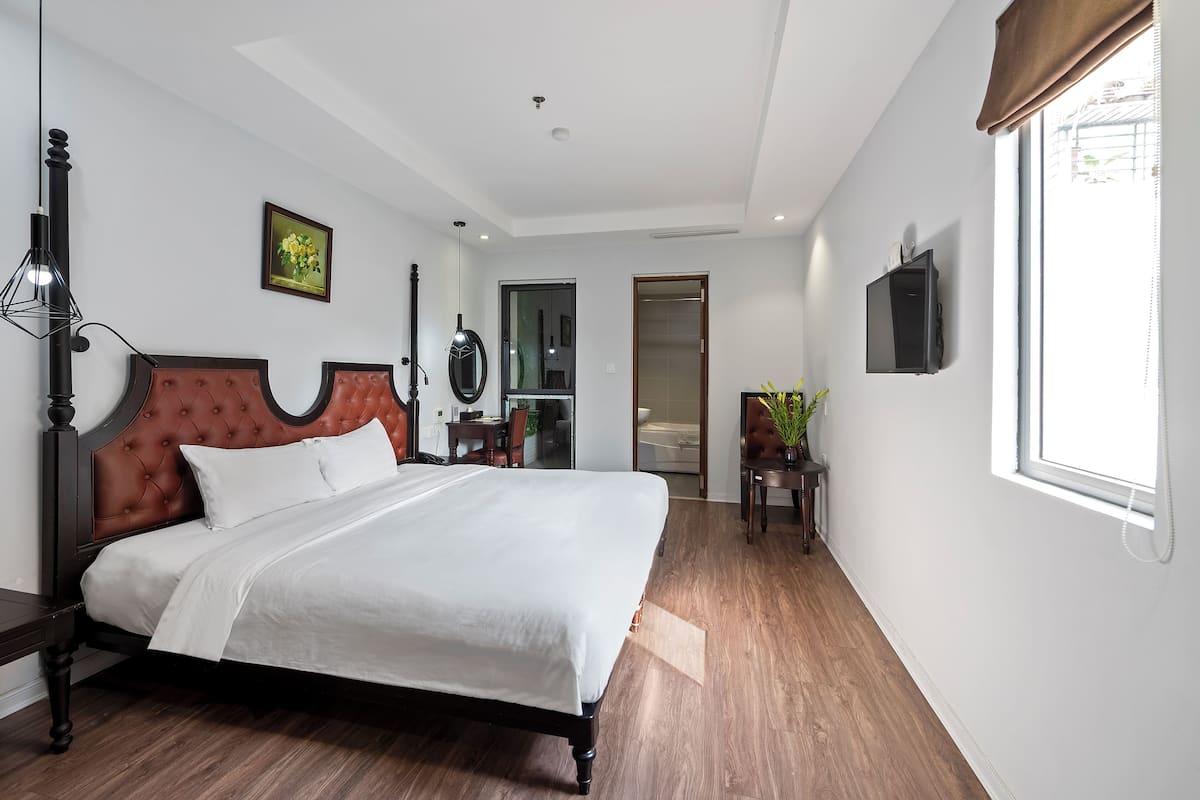 A modern hotel room with a large bed, a wooden floor, a mirror, a TV, and a window with curtains.