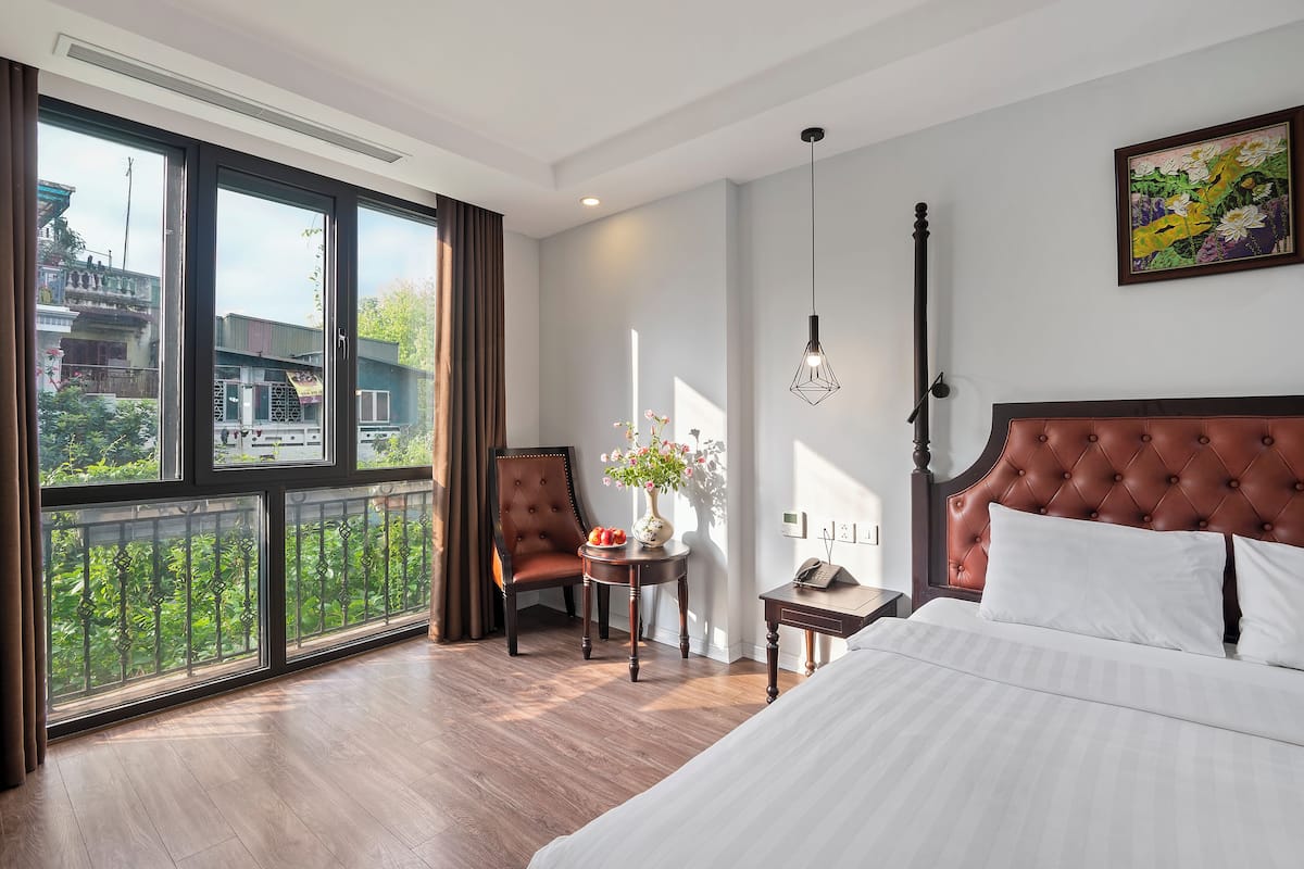 A hotel room with a large window, a bed, a chair, a small table with a vase of flowers, and a painting on the wall.