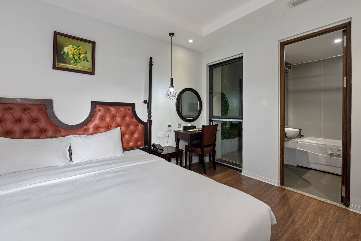 A hotel room with a large bed, a desk with a chair, a mirror, and a bathroom visible through a glass door.