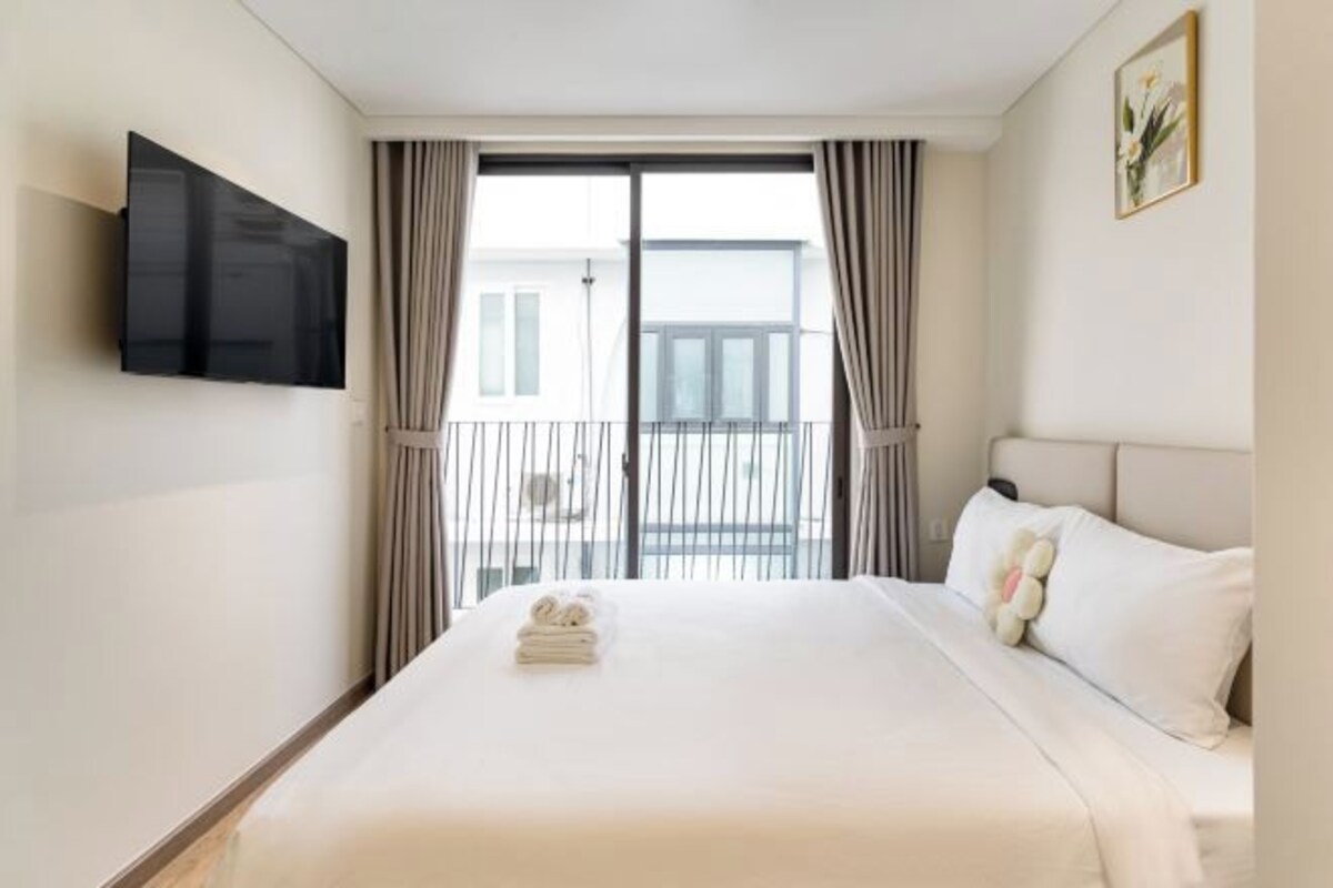 A hotel room with a large bed, a flat-screen TV mounted on the wall, a balcony with curtains, and a painting on the wall.