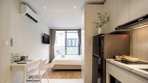 A modern hotel room with a bed, a desk with a chair, a kitchen area with a refrigerator and stove, and a large window with curtains. - The Royal Vista - Phu My Hung (Ho Chi Minh City)