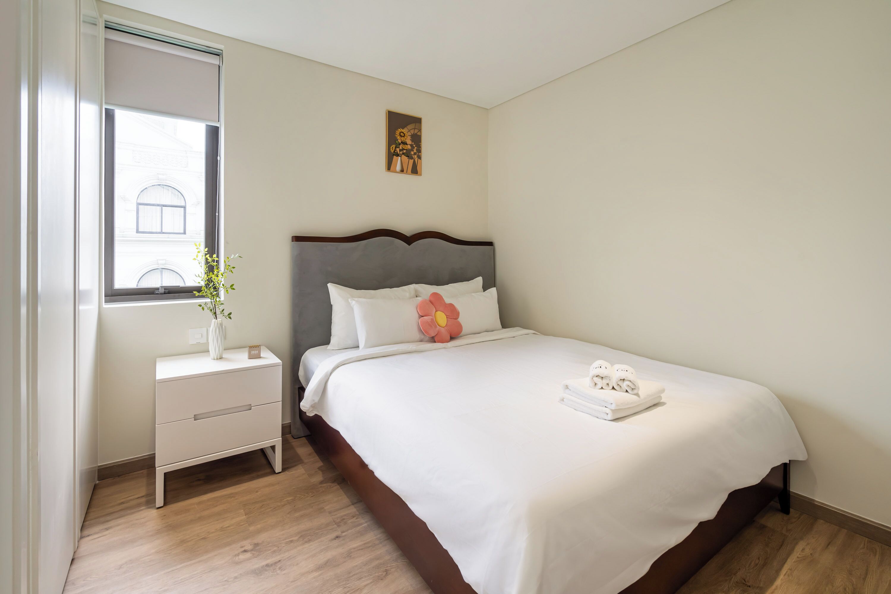 A neatly made bed with white linens, a bedside table with a vase of flowers, and a window with a view of buildings.